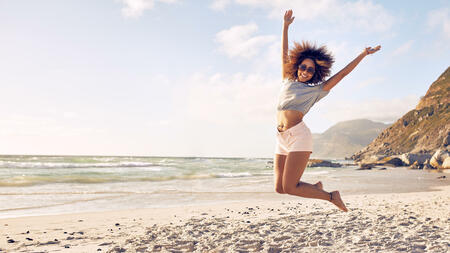 Portrait of beautiful young woman jumping in air at the beach. African female enjoying a summer day at the sea shore.