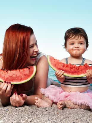 mother with her baby girl lying down on the beach and eating watermelon in summer day.