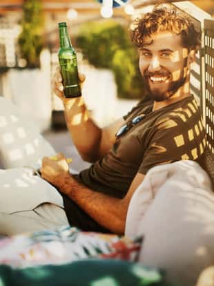 Side view of young man enjoying rooftop party. He's sitting on sofa, smiling, toasting with beer and looking at camera.
