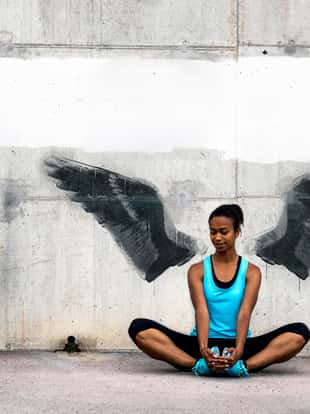 Young woman sitting on the floor, stretching in front of a concrete wall with wings graffiti. The graffiti is not real, the image of wings is my own.