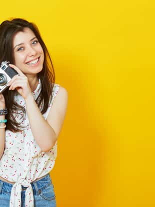 Summer portrait of stylish pretty hipster girl with retro vintage camera posing over yellow background with copy space.