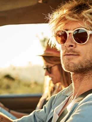 Handsome mid adult man wearing sunglasses by girlfriend in van during sunset. Confident male is driving while female is reading map during road trip. He is having blond hair.