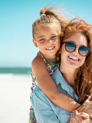 Smiling mother and beautiful daughter having fun on the beach. Portrait of happy woman giving a piggyback ride to cute little girl with copy space. Portrait of happy blonde kid embracing her mom wearing spectacles at beach during summer vacation.