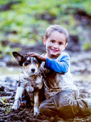 A boy is playing with his dog outside. They are sitting in a mud puddle. The boy looks happy but the dog does not.