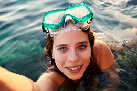 Young woman wearing diving glasses taking selfie looking at camera underwater.