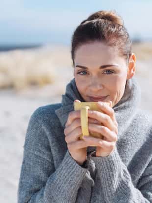 Smiling woman relaxing with coffee at the seaside standing on a sandy beach in a warm winter scarf smiling at the camera