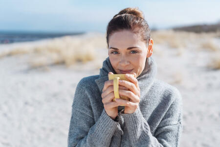 Smiling woman relaxing with coffee at the seaside standing on a sandy beach in a warm winter scarf smiling at the camera