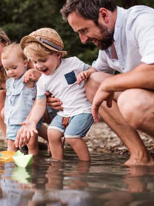 A young family with two toddler children outdoors by the river in summer, playing with paper boats.