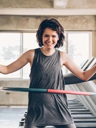 Beautiful young smiling happy Caucasian sporty woman with short hair playing hula hoop inside gym studio with treadmills behind - fun workout fitness portrait concept