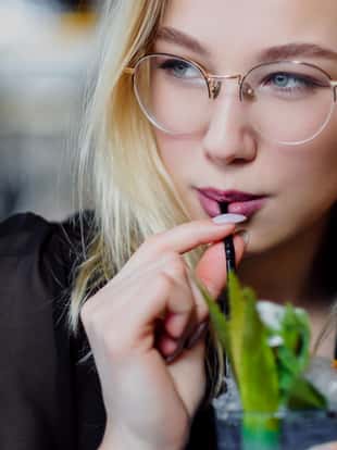 A young beautiful blond woman with glasses sits at the bar in a yellow interior. Drinks alcoholic cocktail with a straw and green mint. Night life. and rest at a party.