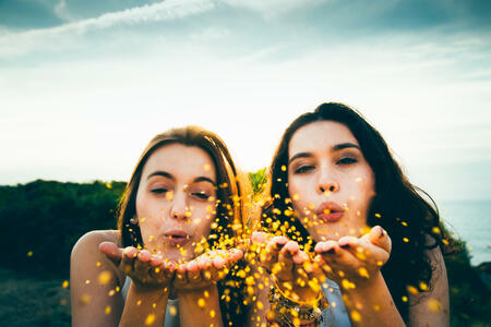 Funny female friends blowing golden glitter over a cliff at dusk