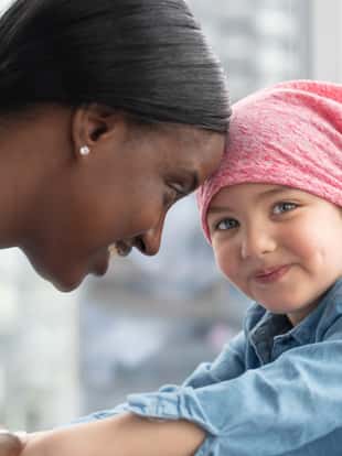 A cute elementary age girl with cancer is wearing a pink scarf on her head. She is at a medical appointment. The female doctor of African descent is holding the child's hands, providing comfort and support. The child is smiling at the camera.