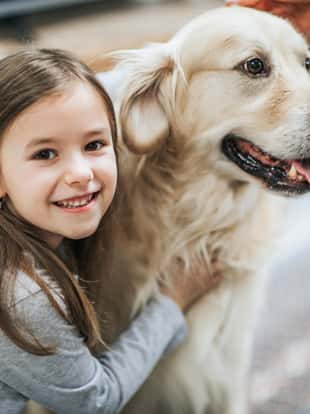 Cute little girl enjoying while embracing her dog at home and looking at camera.
